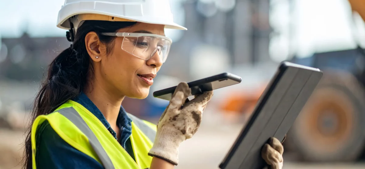 Woman in safety gear using mobile FLHA tools with voice-to-text on a construction site.
