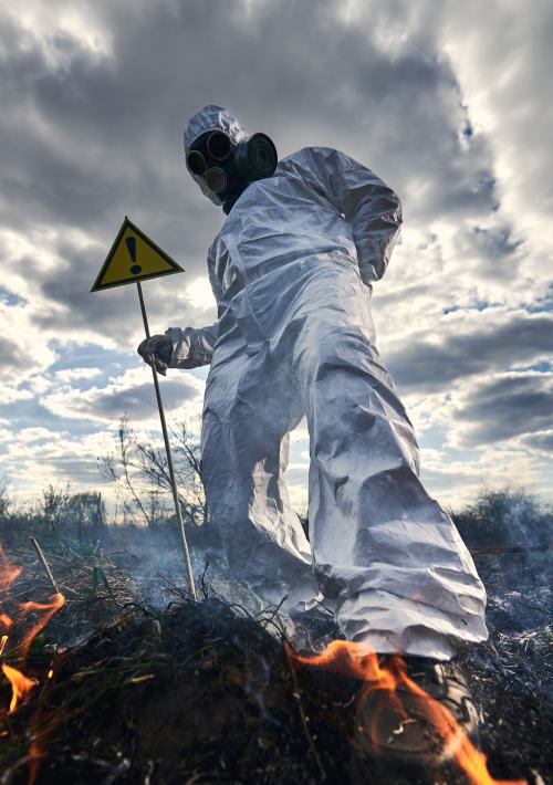 Fireman ecologist fighting fire in field. Man in protective radiation suit and gas mask near burning grass with smoke, holding warning sign with exclamation mark. Natural disaster concept.