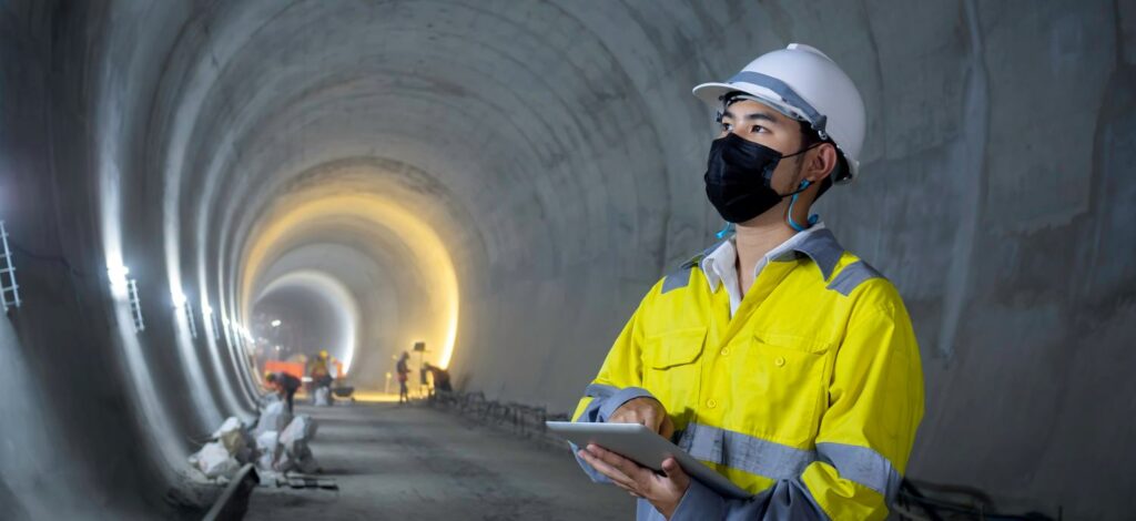 Young Asian tunnel engineering wearing high visibility jacket, face mask and white safety helmet working and using digital tablet in dark railway tunnel construction site area with copy space.