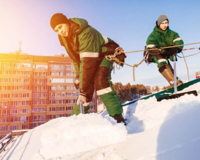 Snow cleaning. Team of male workers clean roof of building from snow with shovels in securing belts of mantra.