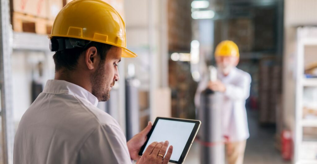 Picture of young focused male manager standing in factory and holding tablet. Dressed in white coat with helmet on his head.