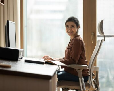 Modern day worker. Portrait of happy biracial business woman freelancer sit by computer at comfy workplace at corporate workspace or at home. Smiling young indian lady office employee look at camera