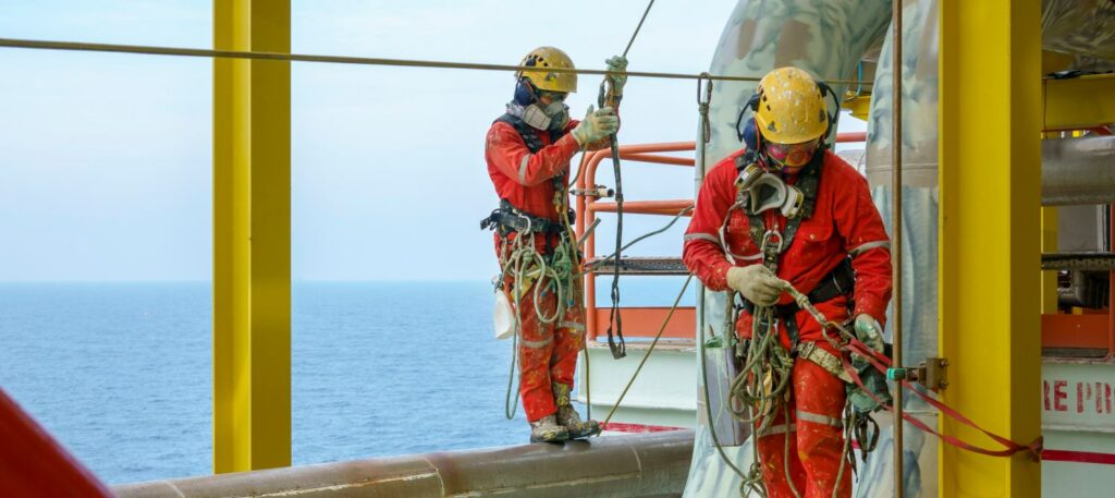an example of a good EHS system, two safe specialists working at heights. A group of abseilers wearing red coverall and Personal Protective Equipment (PPE) such as hard hat, harness, hand protection and eye protection standing on the pipeline arranging their rope access with background open sea.