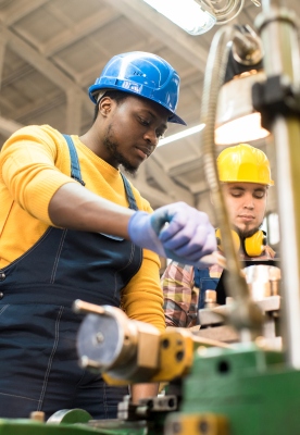 Multi-ethnic team of workers wearing overalls and protective helmets using lathe in order to machine work piece, interior of spacious production department on background