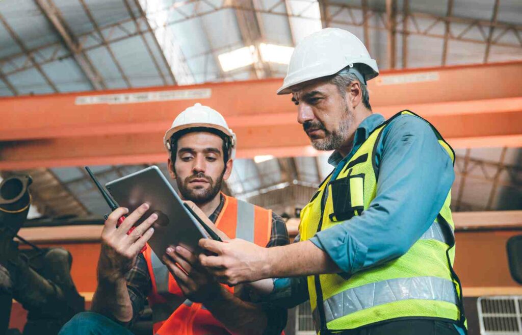 Two senior and young male engineers in safety vest and jacket with hardhat and helmet working in warehouse and factory on a machine while fixing and inspecting equipment