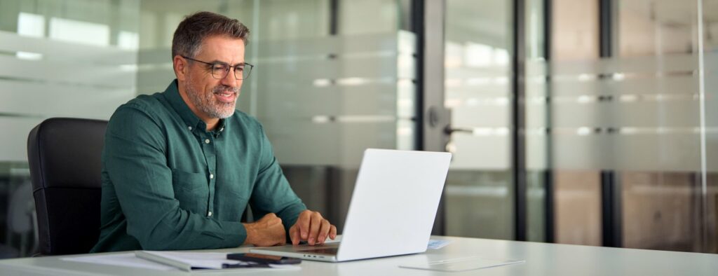 Smiling busy older professional business man working on laptop sitting at desk while taking a virtual proctored online course