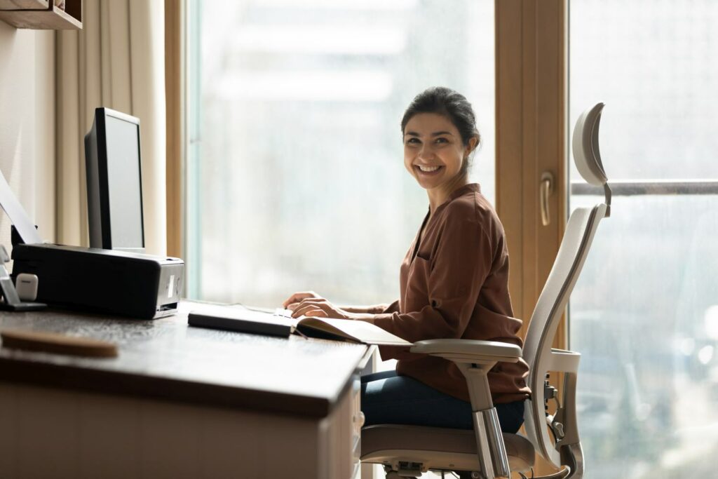 Modern day worker. Portrait of happy biracial business woman freelancer sit by computer at comfy workplace at corporate workspace or at home.