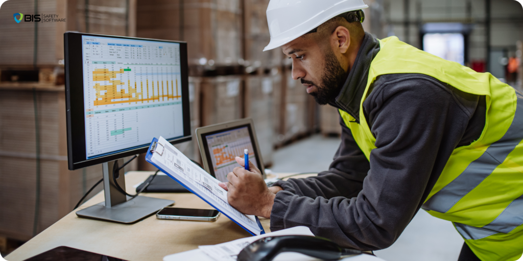 Construction site manager reviews documents while analyzing data on a computer monitor in a warehouse.