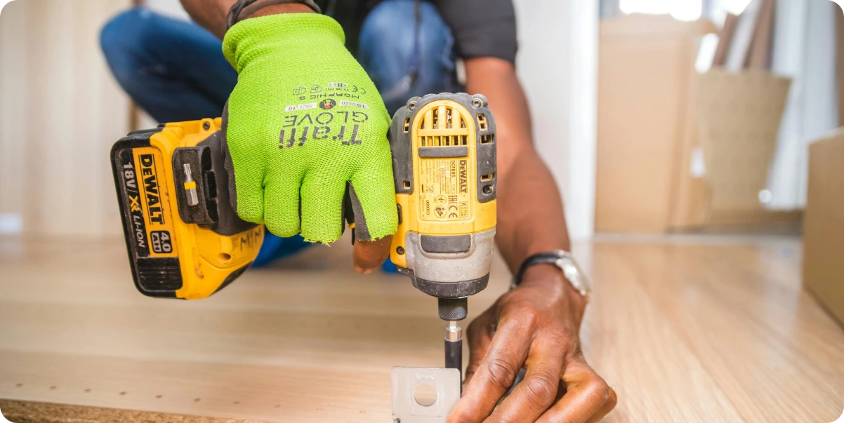A worker wearing a bright green safety glove uses a power drill to fasten a metal bracket to a wooden board, symbolizing how everyday workers quietly reshape safety practices through hands-on choices and small acts of responsibility.